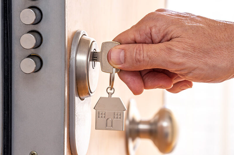 Eine Männerhand hält einen Schlüssel mit einem Anhänger in Form eines Hauses fest, der im Schloss einer Eingangstür mit einbruchsicherer Dreipunkt-Verriegelung steckt.